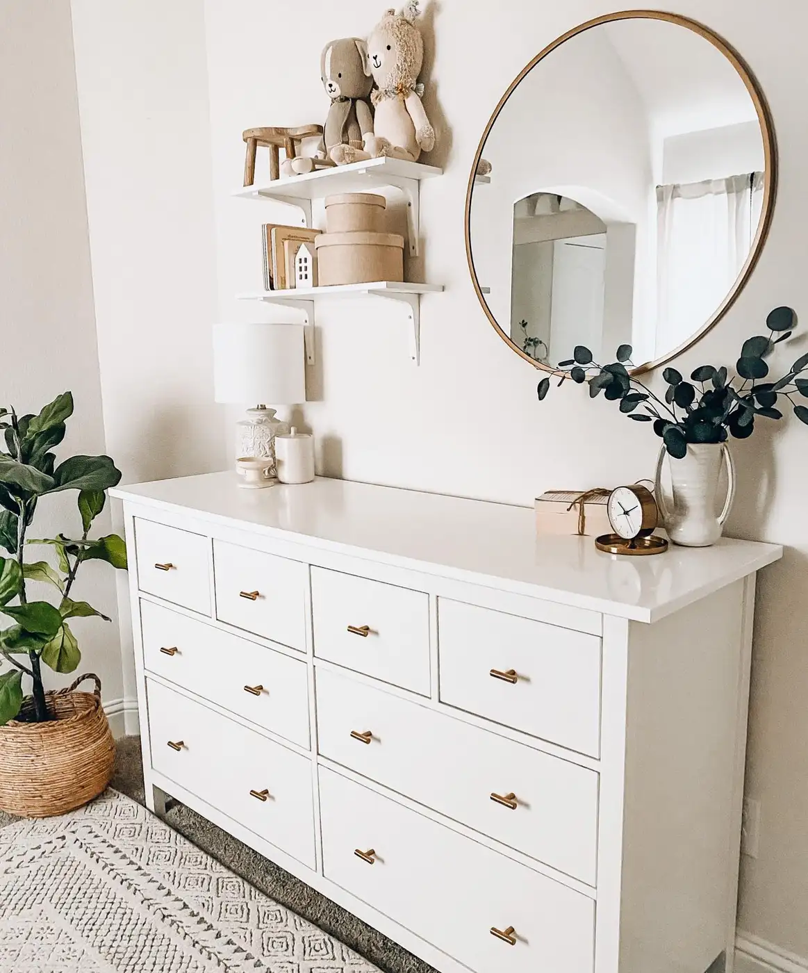 White dresser with round mirror, table lamp, leafy branch, and small grouped accessories