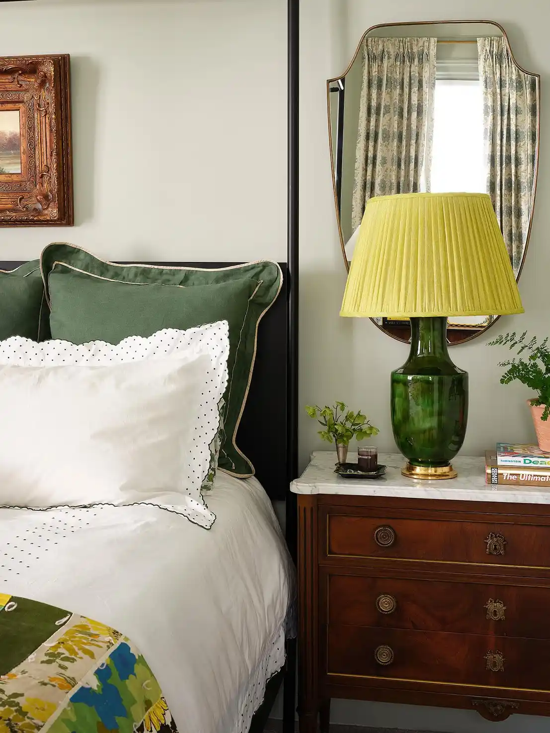 Nightstand with green glass lamp, yellow pleated shade, small plant, books, and wall mirror