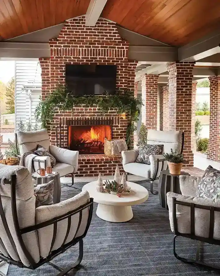 Large covered porch with a brick fireplace, wood ceiling, pale upholstered seating, light rug, and plants.