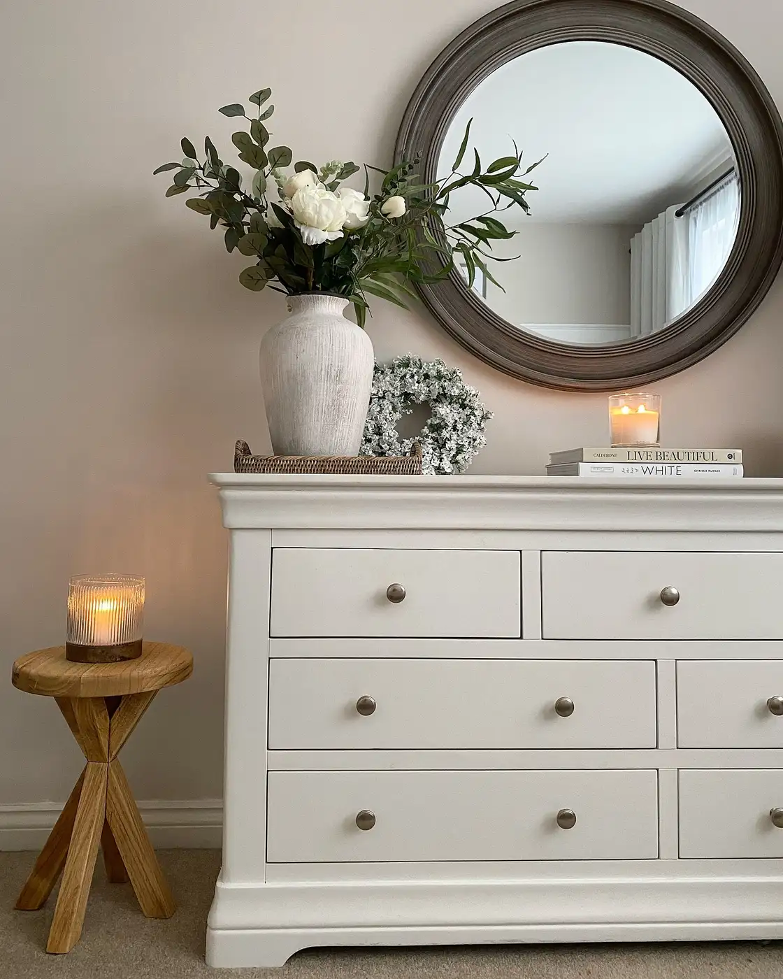 White dresser beneath round mirror with tall white flowers, stacked books, and simple decorative accents