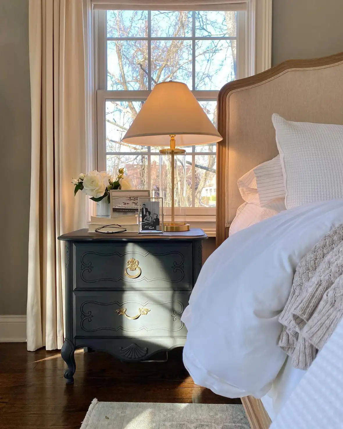 Small dark nightstand with lamp, tiny flower vase, and framed photo beside the bed