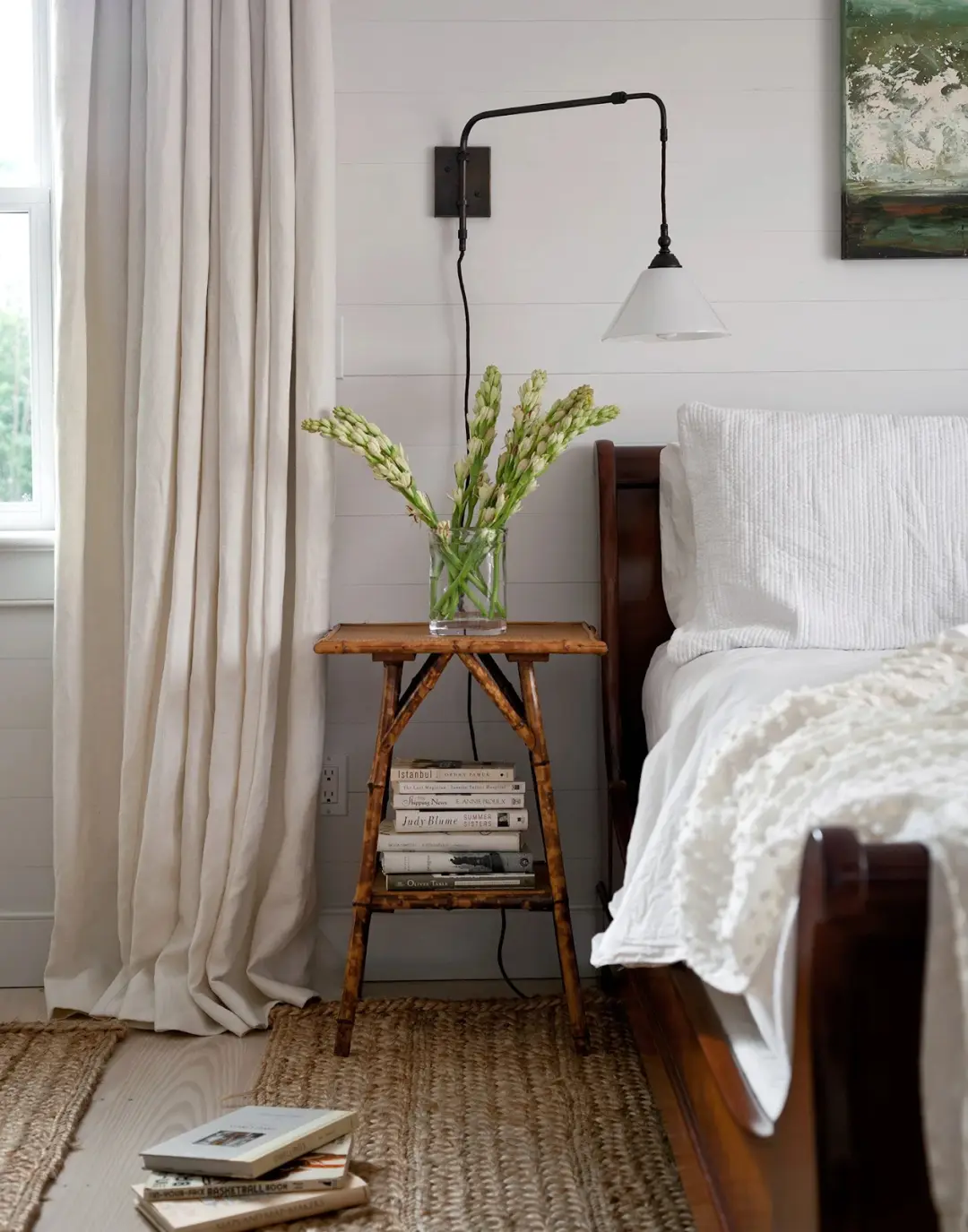 Slim wooden nightstand with wall-mounted light, flower vase, and books on lower shelf