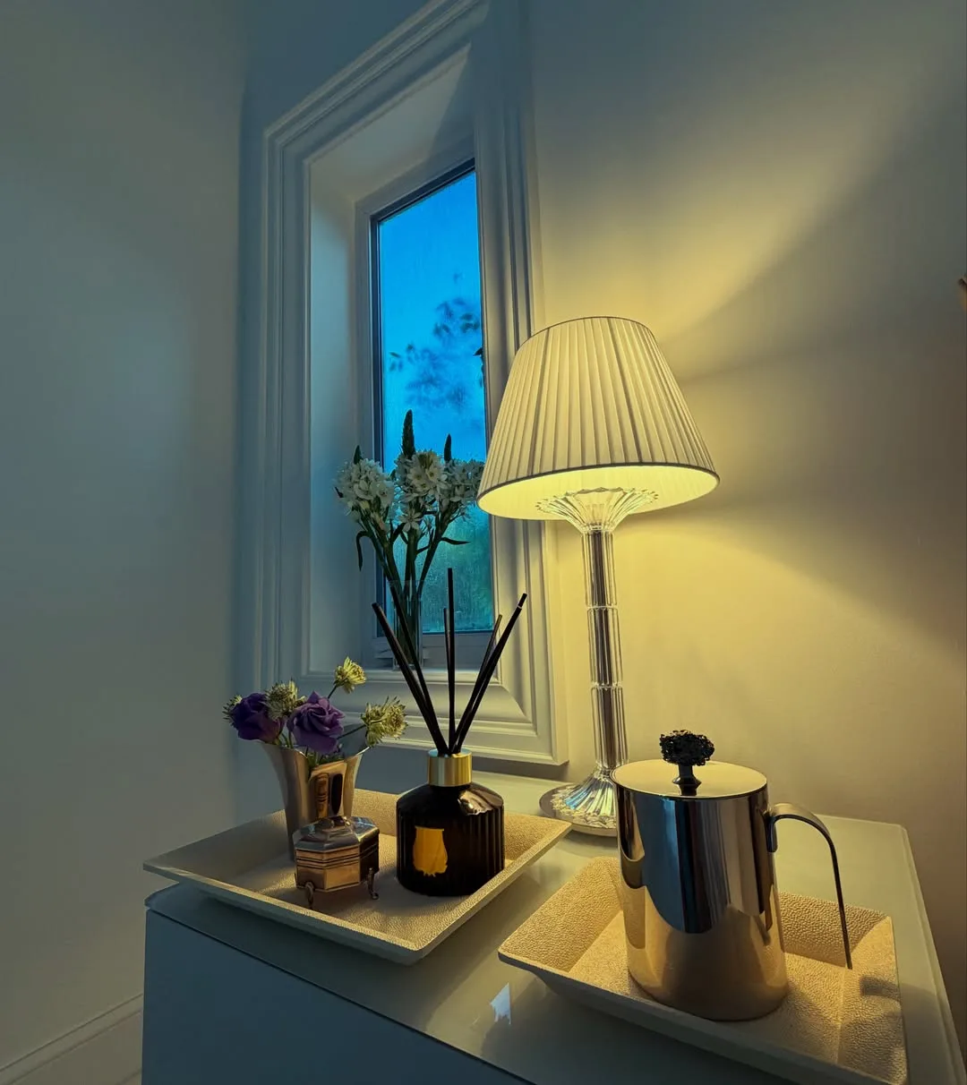 Glossy nightstand with lamp, tray of candles and flowers, diffuser, and metal pitcher