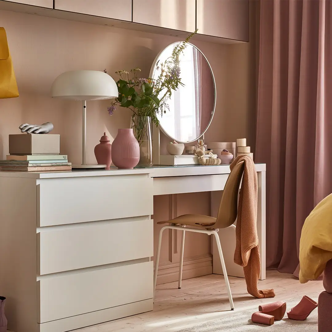 Long pink-toned dresser arranged as a vanity with round mirror, chair, lamp, books, and flowers