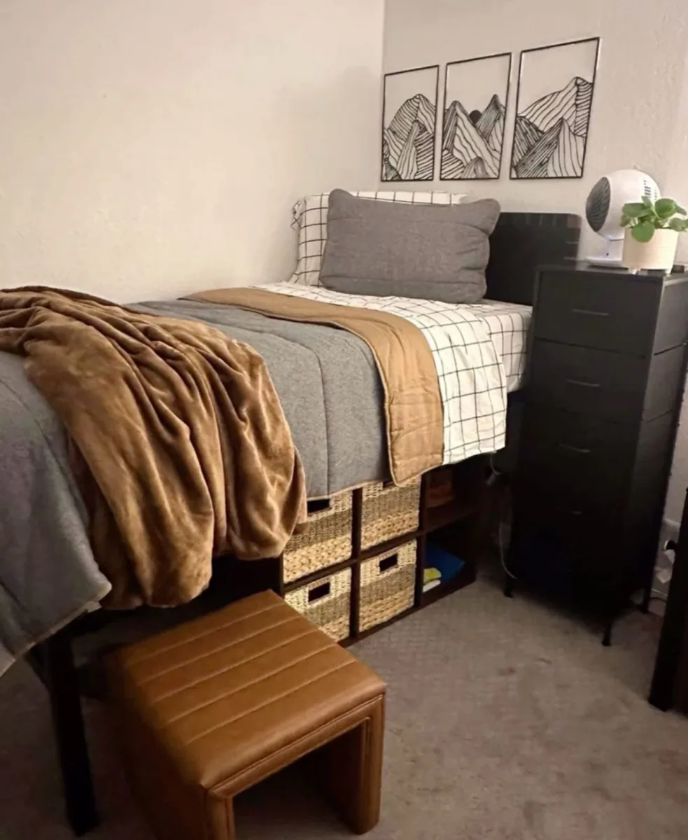 Gray dorm bed styled with a camel brown throw, black furniture, and a small wooden stool