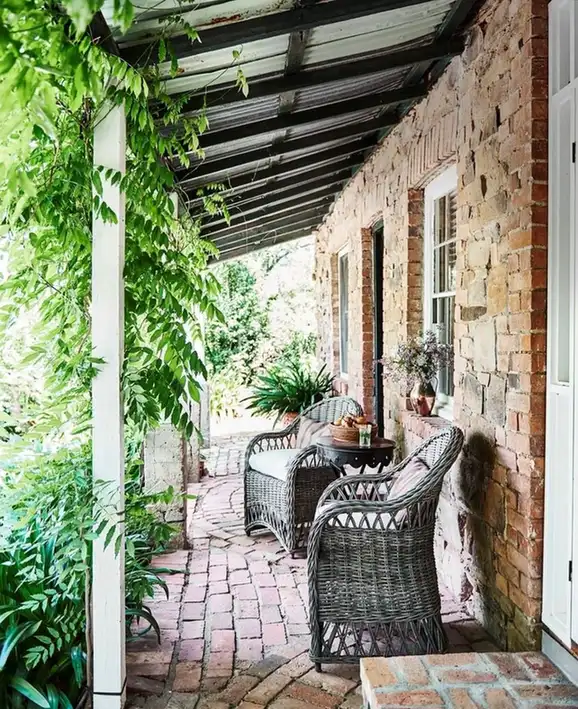 Narrow covered porch with black chairs, brick floor, stone wall, and climbing greenery