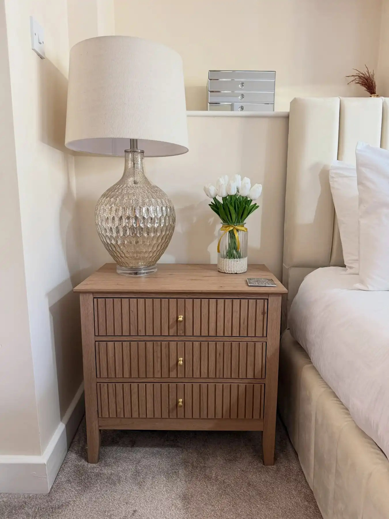 Wood nightstand with large glass lamp, small vase of white flowers, and neutral bedding