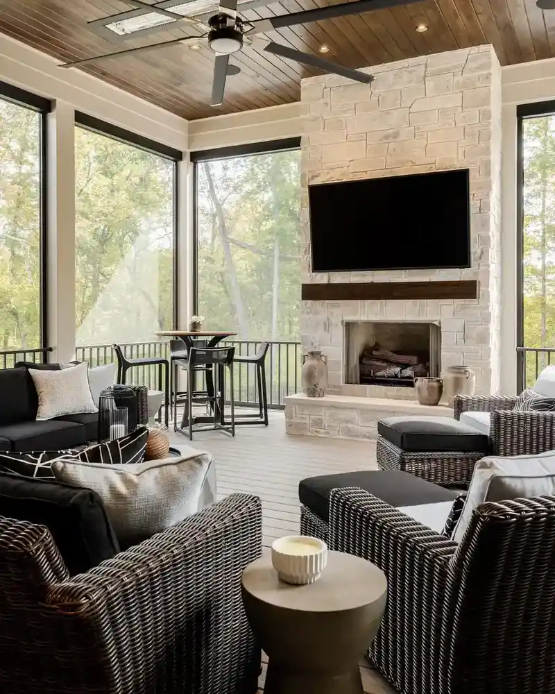 Screened porch with a TV mounted above a stone fireplace, dark seating, wood ceiling, and windows.