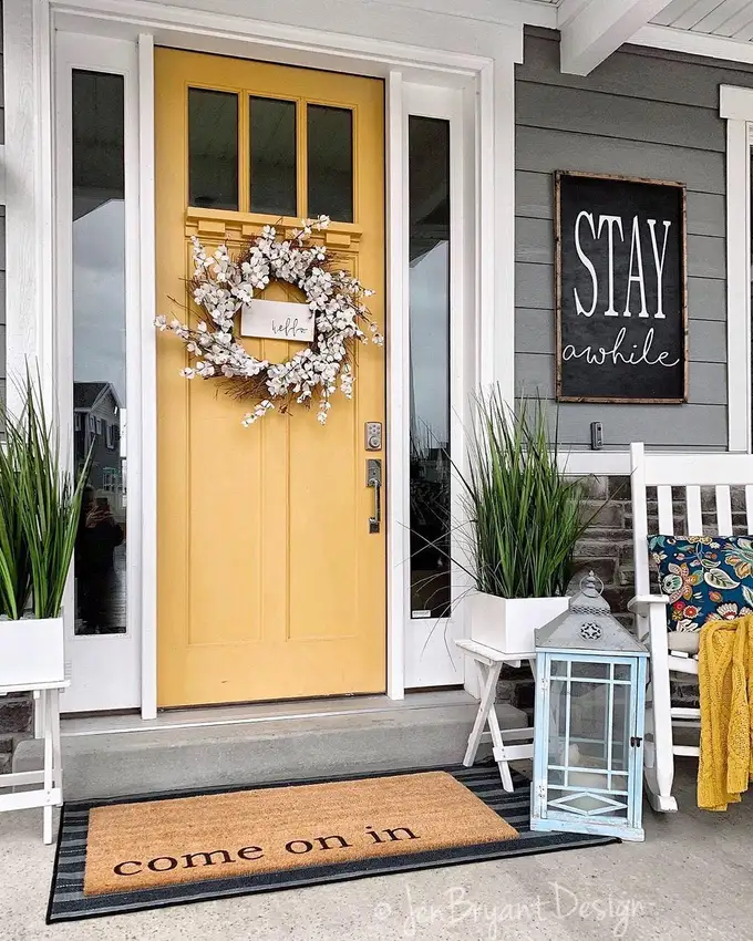 Yellow front door with a white floral wreath, black shutters, white trim, and green plants