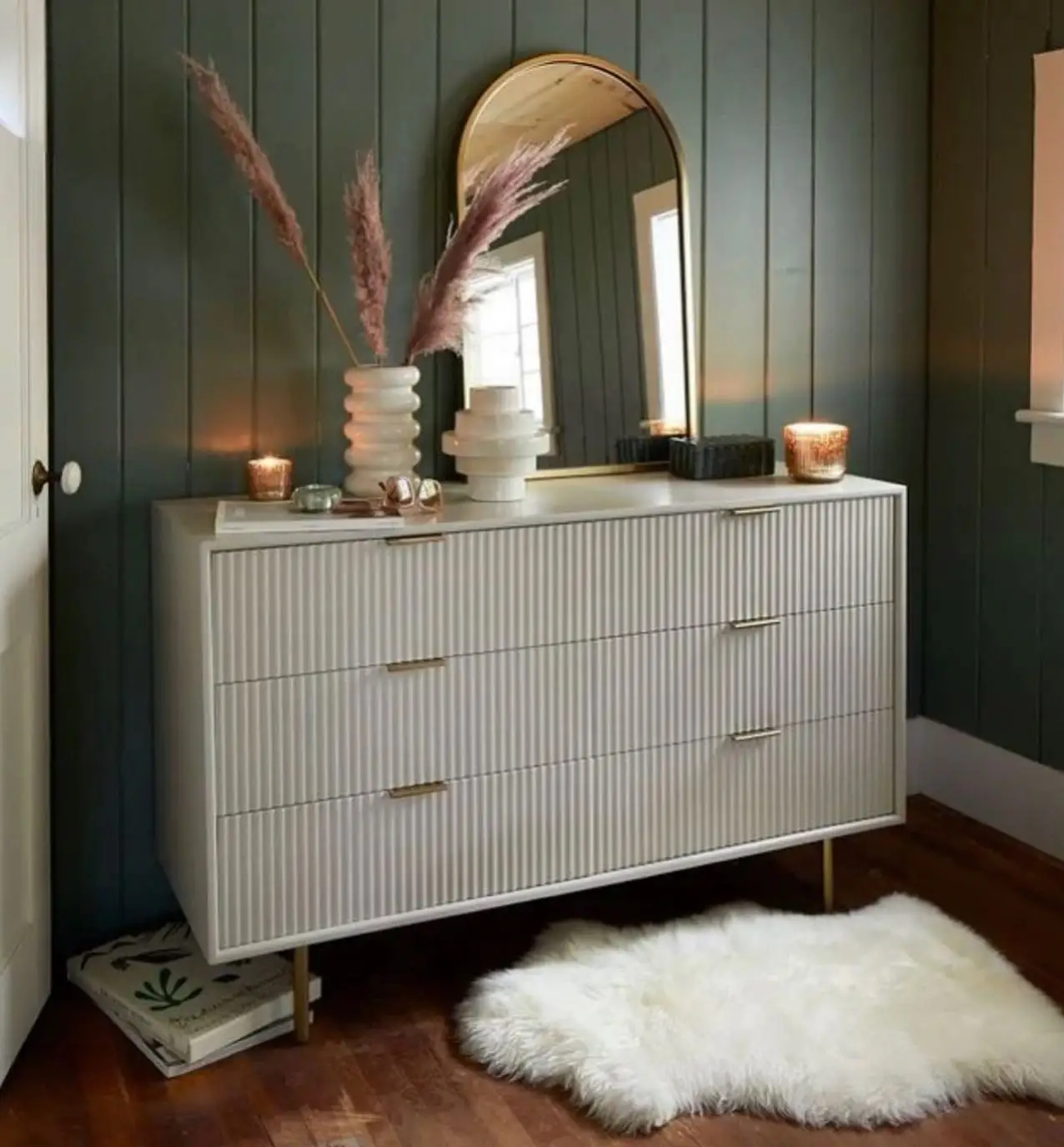 Light dresser against deep green paneled wall with round mirror, dried stems, candles, and sculptural vases