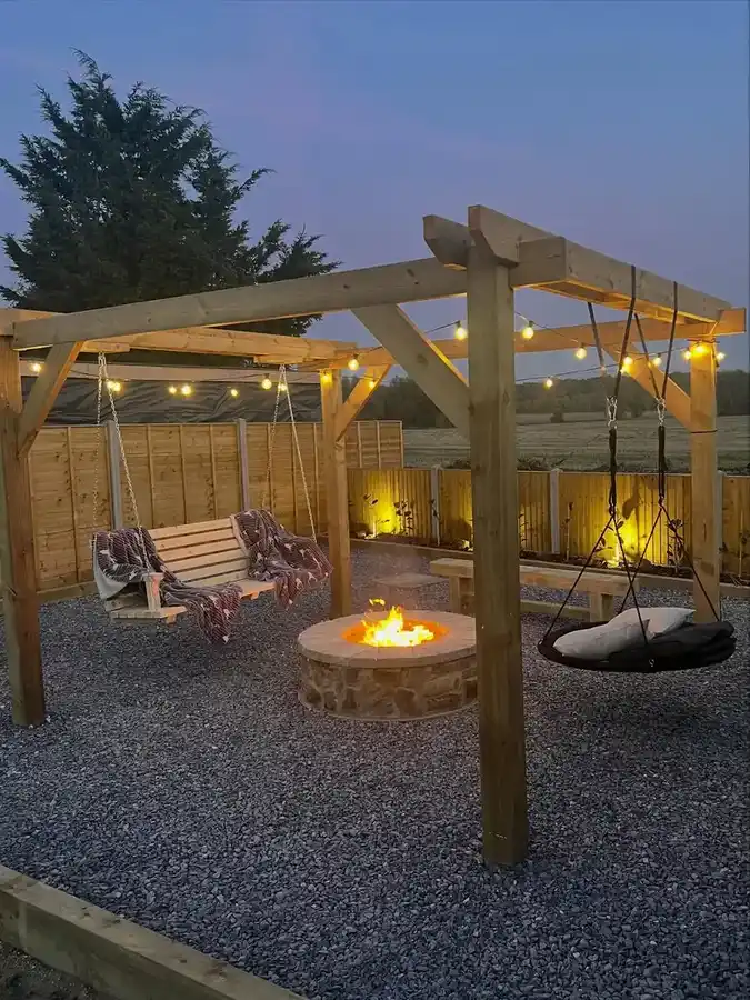 Backyard fire pit with wooden pergola, gravel seating area, outdoor chairs, and string lights