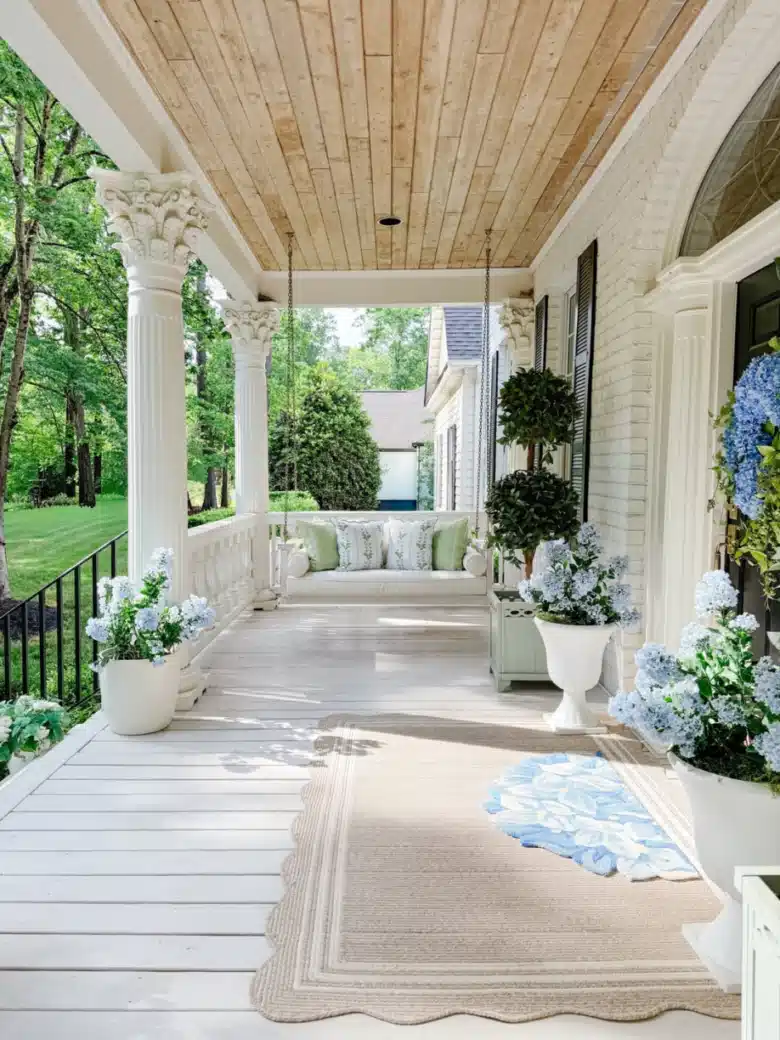 Bright farmhouse porch with white furniture, pale flooring, a natural wood ceiling, and green potted plants