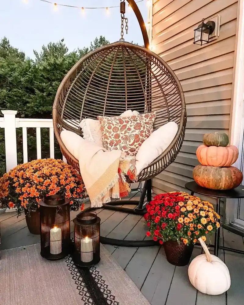Covered porch corner with a hanging wicker egg chair, cushions, pumpkins, flowers, and lanterns.