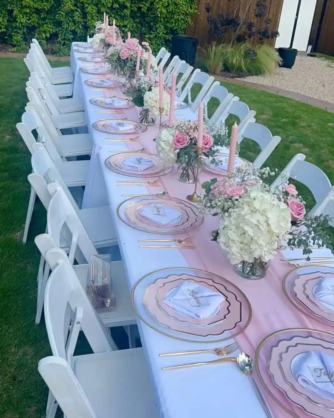 Outdoor garden party table with pink napkins, pale tableware, white chairs, and floral centerpieces