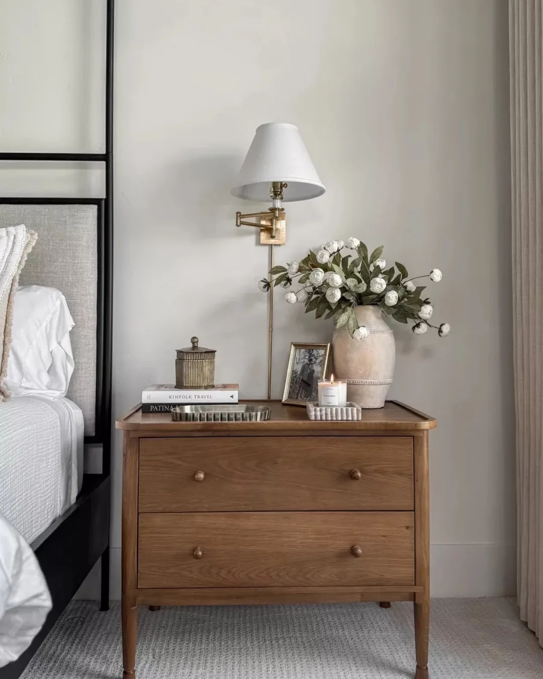 Nightstand with wall sconce, white flowers, framed photo, books, and small bedside accessories