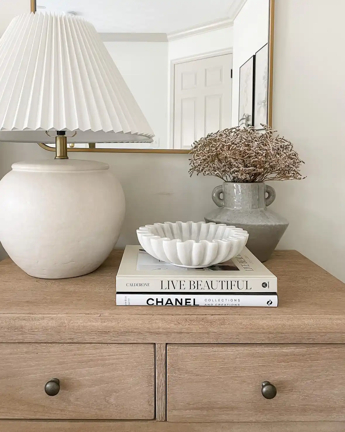 Neutral nightstand with large rounded lamp, handled vase, dried stems, fluted bowl, and books
