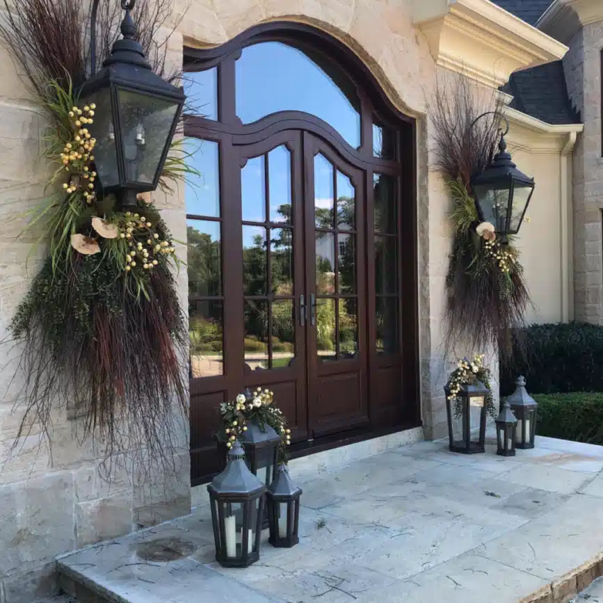 Rustic farmhouse entrance with a tall arched wooden door, stone surround, large planters, and lanterns