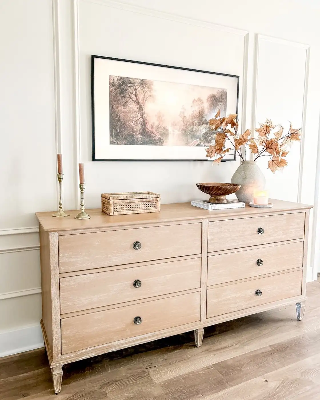 Light wood dresser with dried brown branches, woven storage box, wooden bowl, and framed landscape art