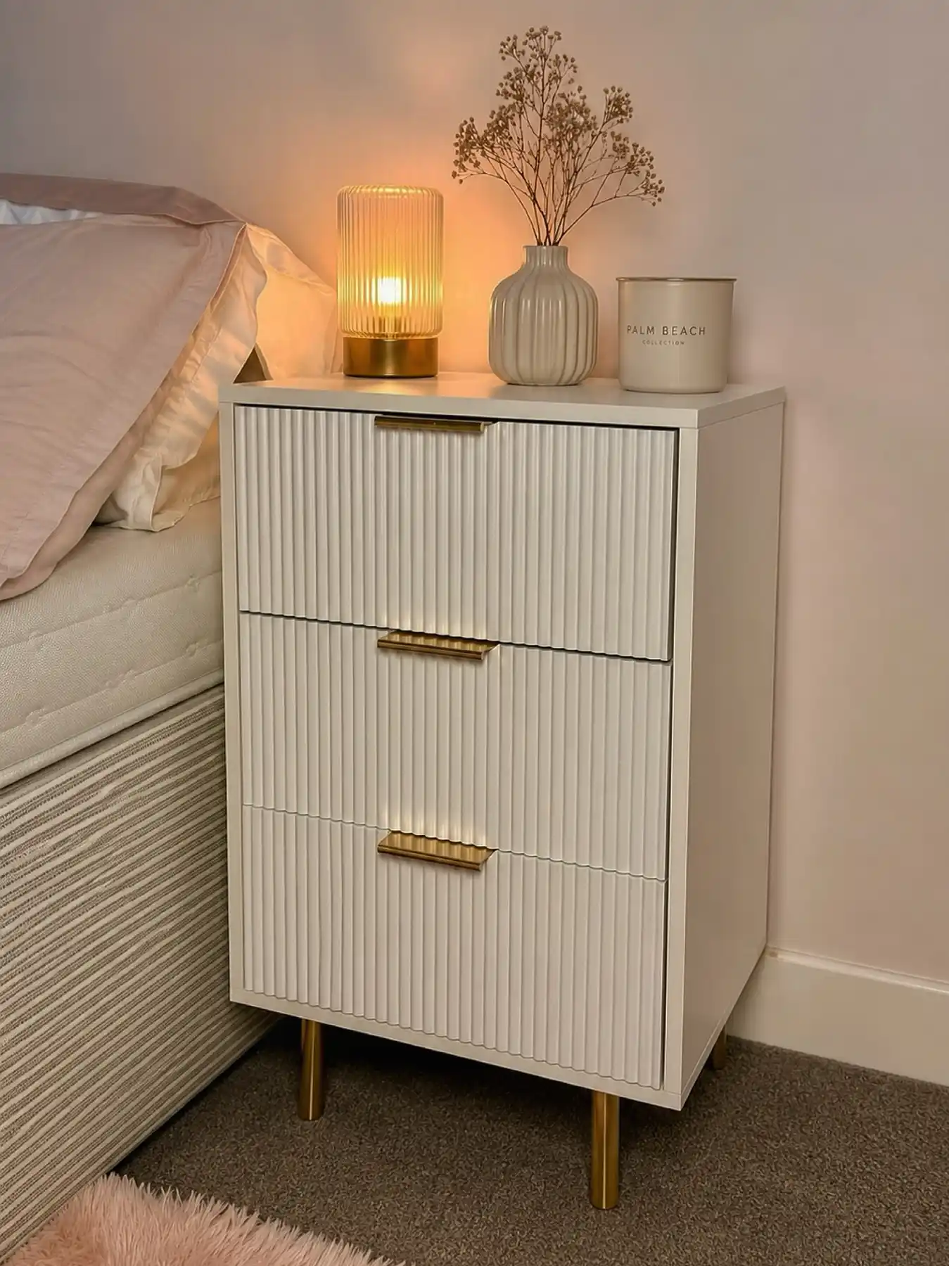 Tall narrow nightstand with three drawers, lamp, slim flower vase, candle, and gold hardware