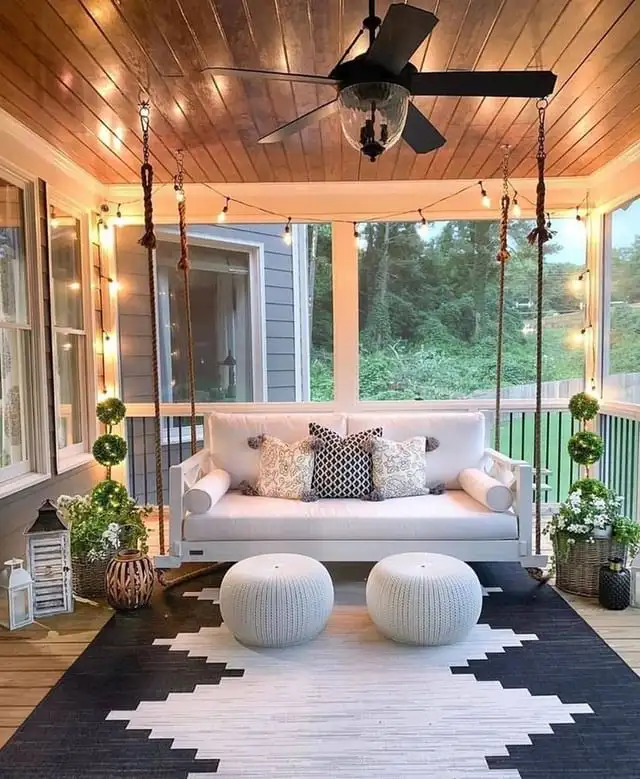 Screened porch with warm wood ceiling, dark ceiling fan, pale sofa, round ottomans, patterned rug, and plants.