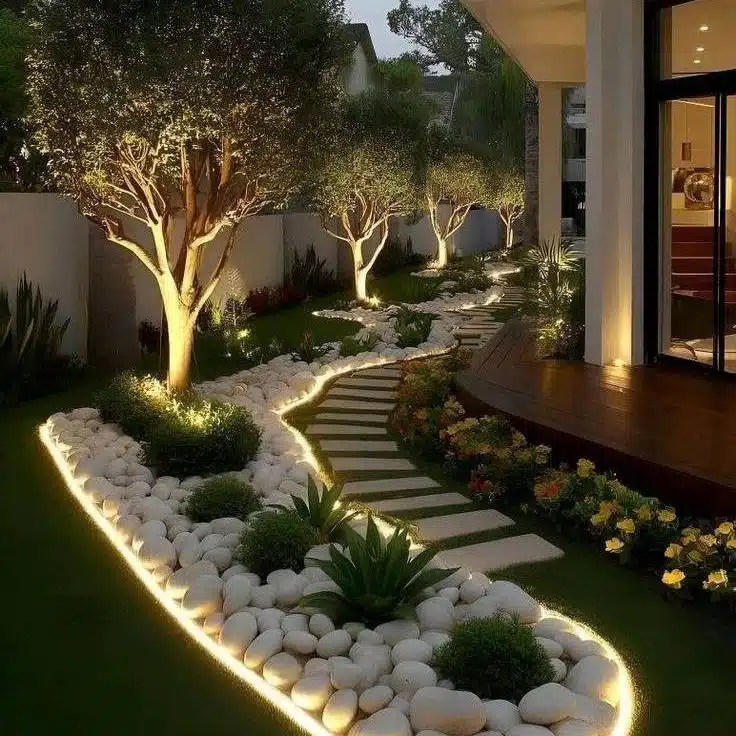 Outdoor uplighting illuminating tree trunks and branches beside a garden bed with pale decorative stones.