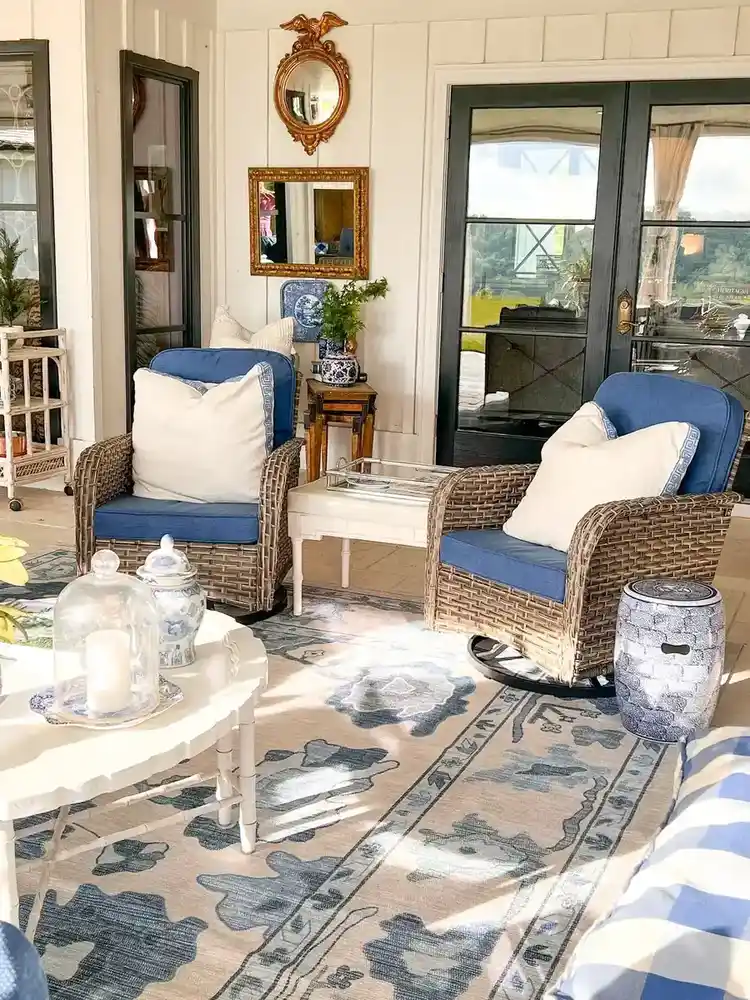 Bright covered porch with pale seating, blue cushions, patterned rug, small table, and large windows.