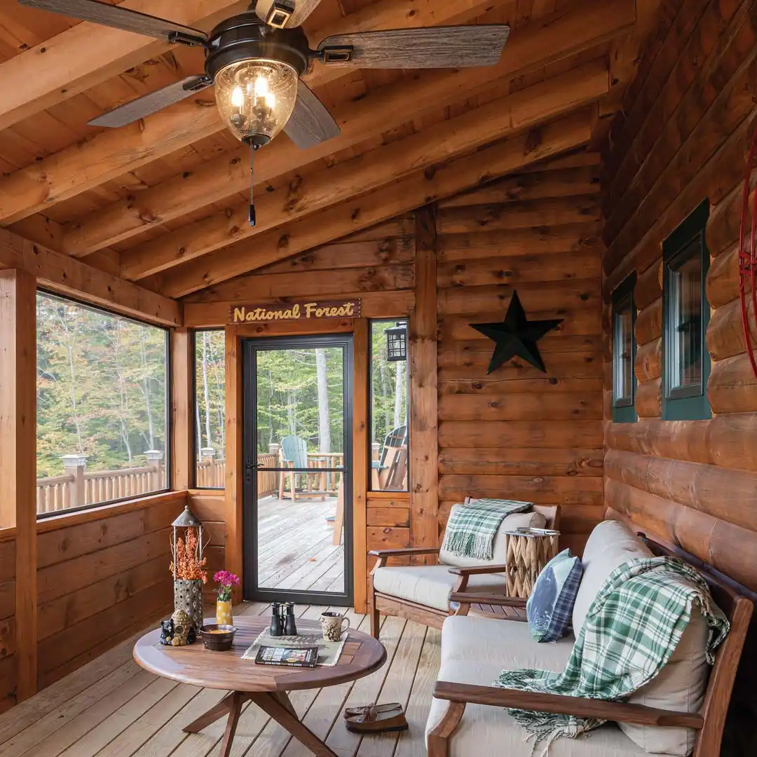 Enclosed rustic porch with wood walls and ceiling, wicker seating, patterned cushions, and plants