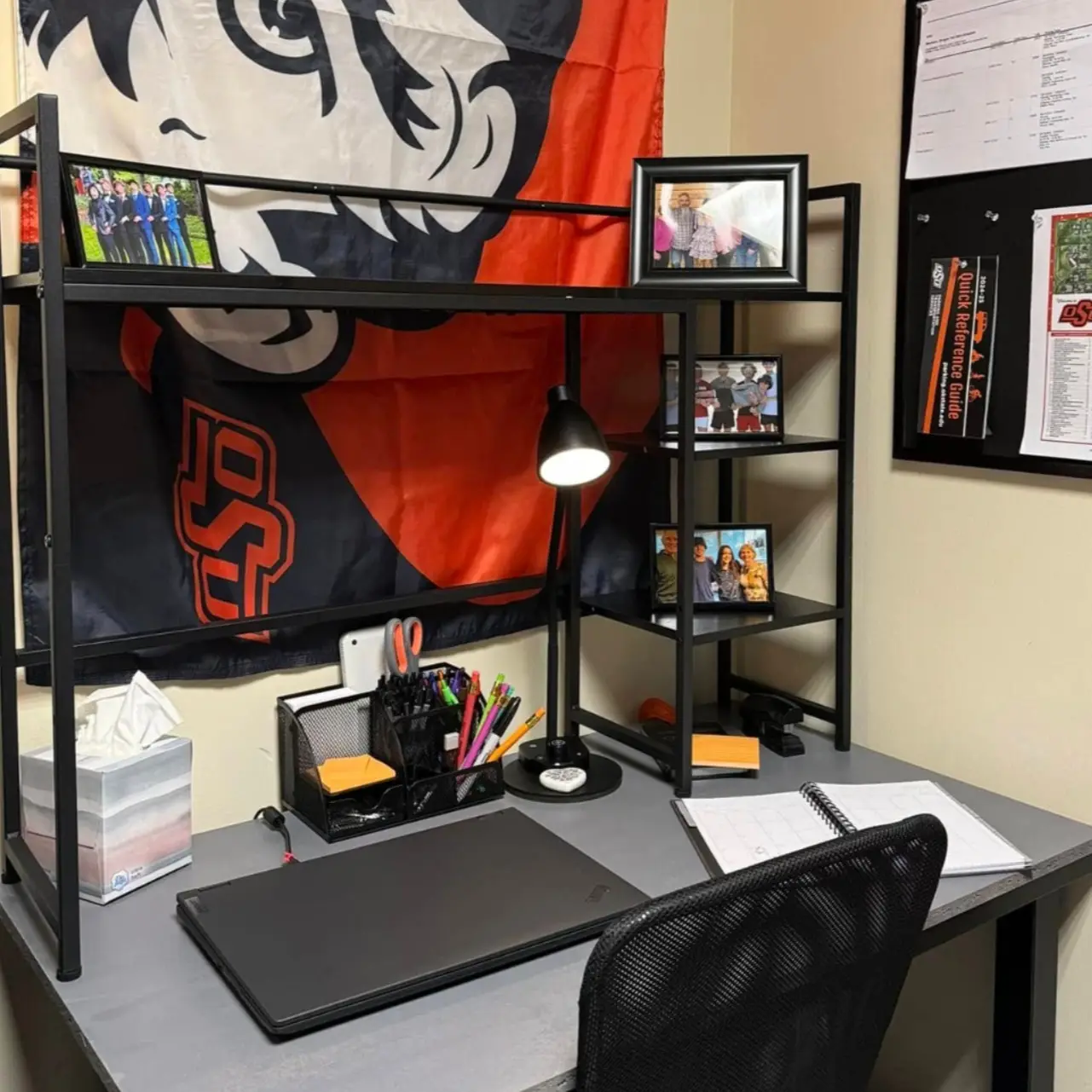 Black framed dorm desk with shelving, large wall flag, lamp, and simple accessories