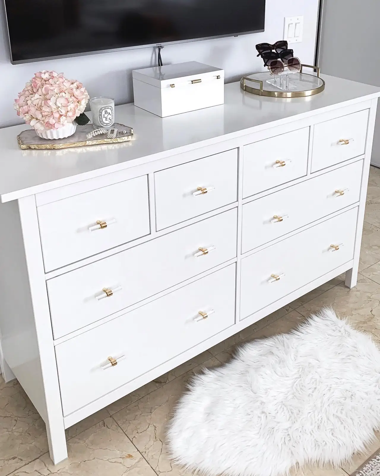 Wood dresser beneath wall-mounted TV with low flowers, storage box, and small round tray