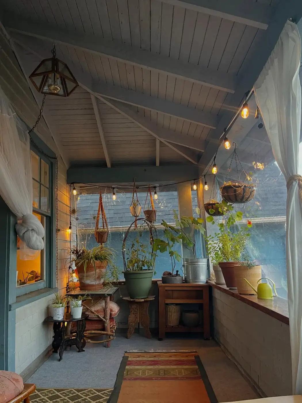 Cozy outdoor dining porch with a long table, string lights, greenery, and evening lighting.