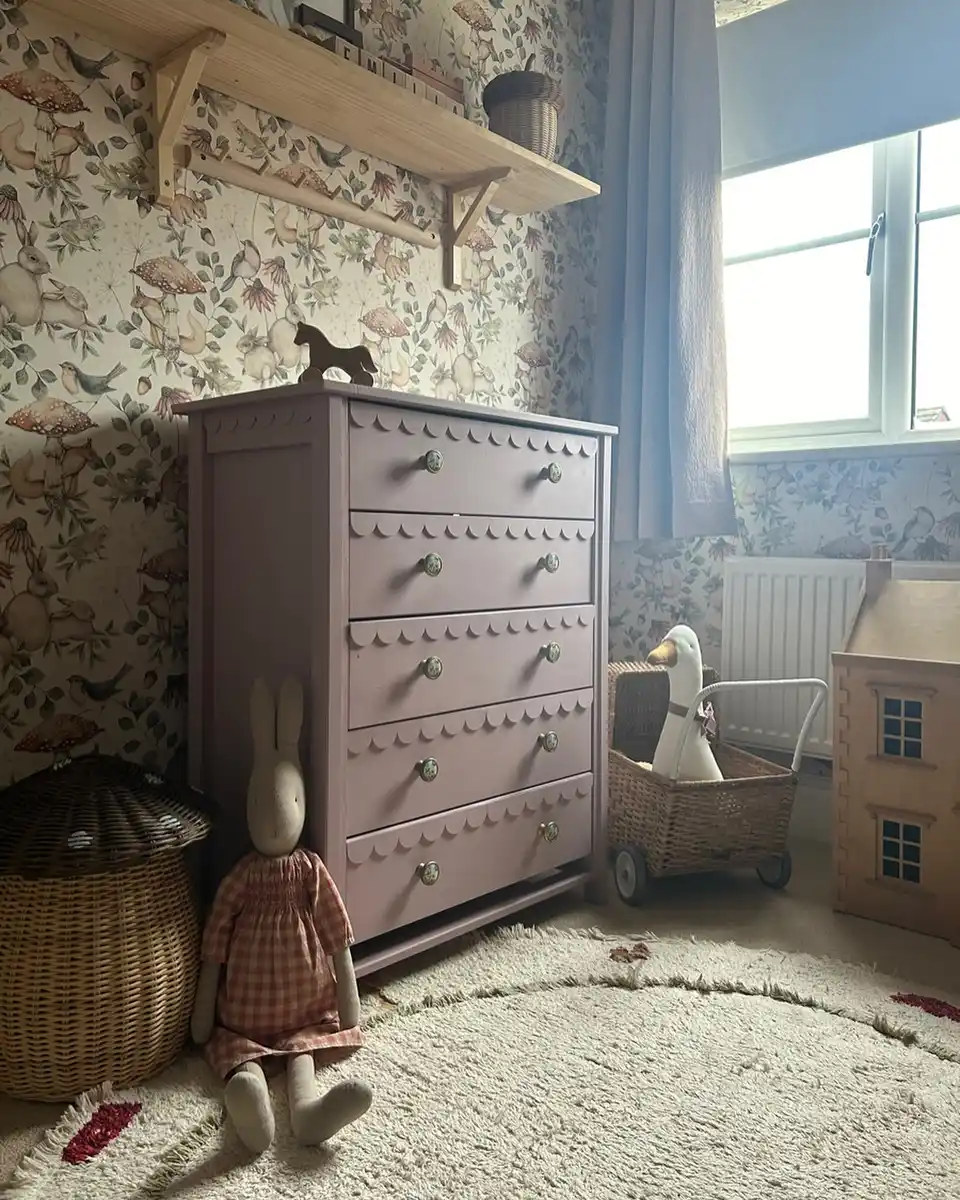 Tall nursery dresser against floral wallpaper with shelves, baskets, toys, and patterned rug nearby