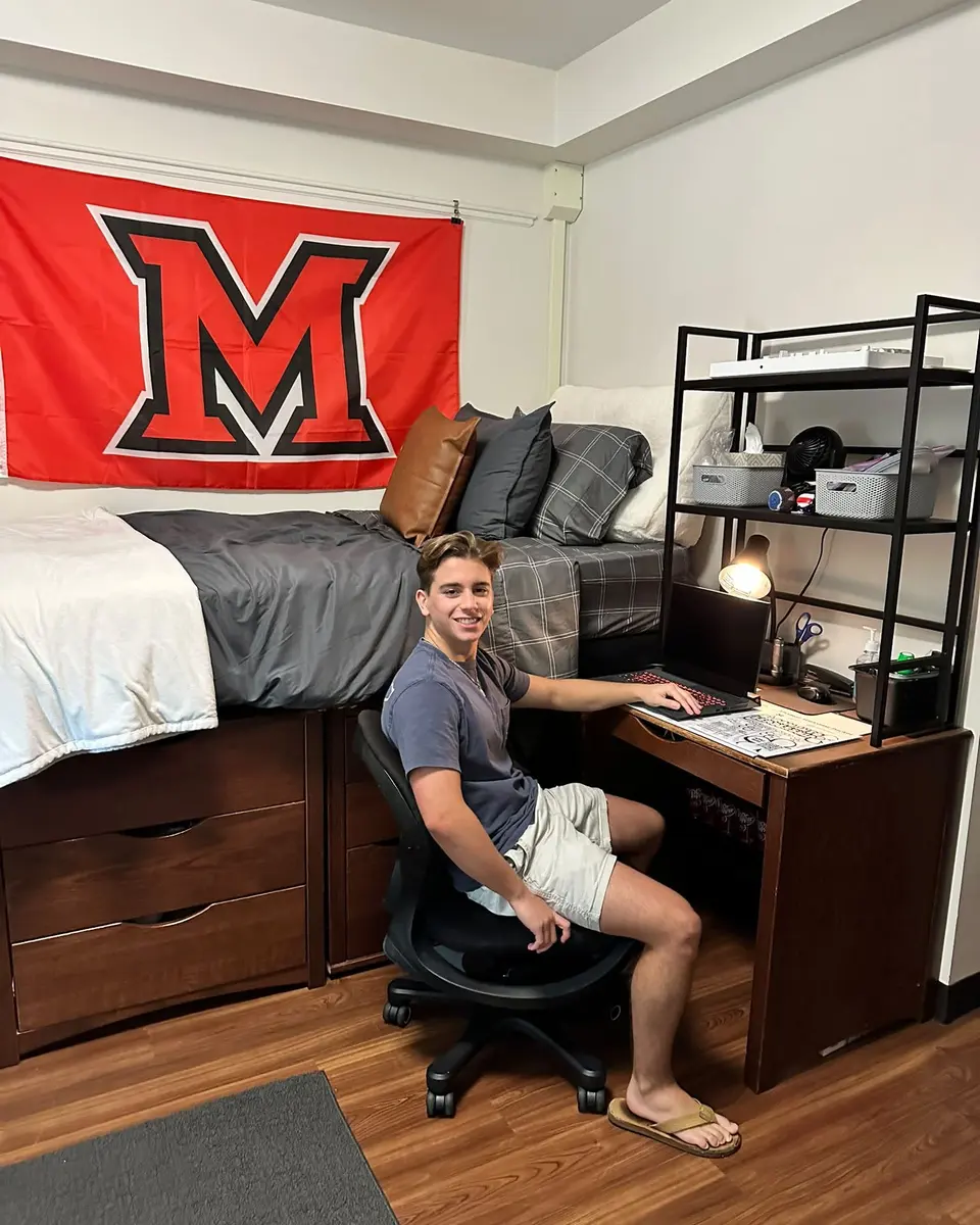 Compact dorm room with a desk beside a raised bed and tall open shelving above the workspace