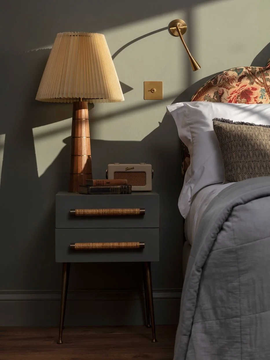 Nightstand with tall pleated lamp, adjustable wall light, books, and small bedside clock