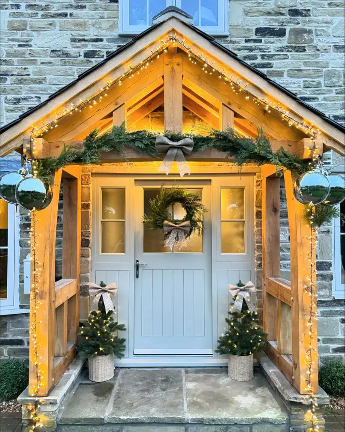 Light blue front door beneath a peaked wooden awning with warm lights, greenery, and potted plants
