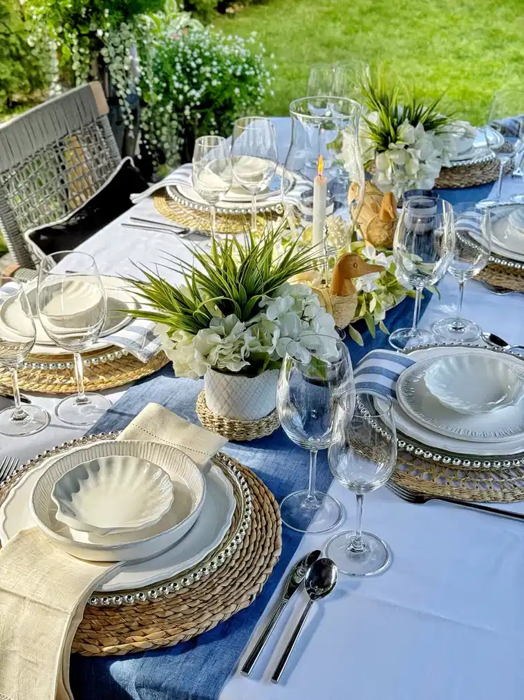 Outdoor dining table with blue and white plates, pale linens, woven placemats, and white flowers
