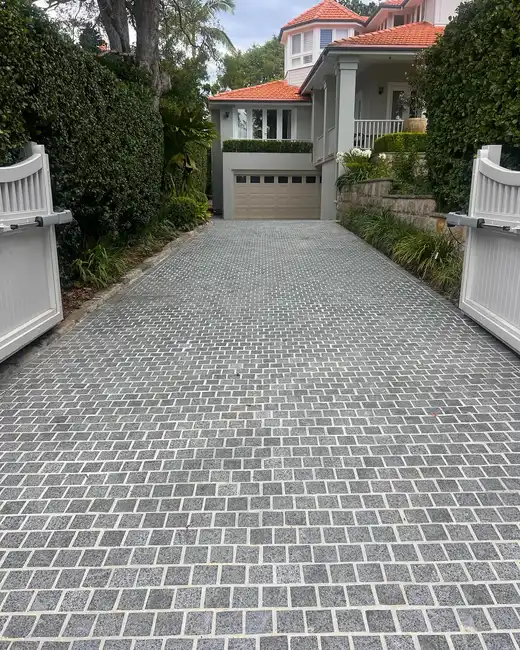Long driveway covered with small gray pavers and bordered by tall hedges and walls
