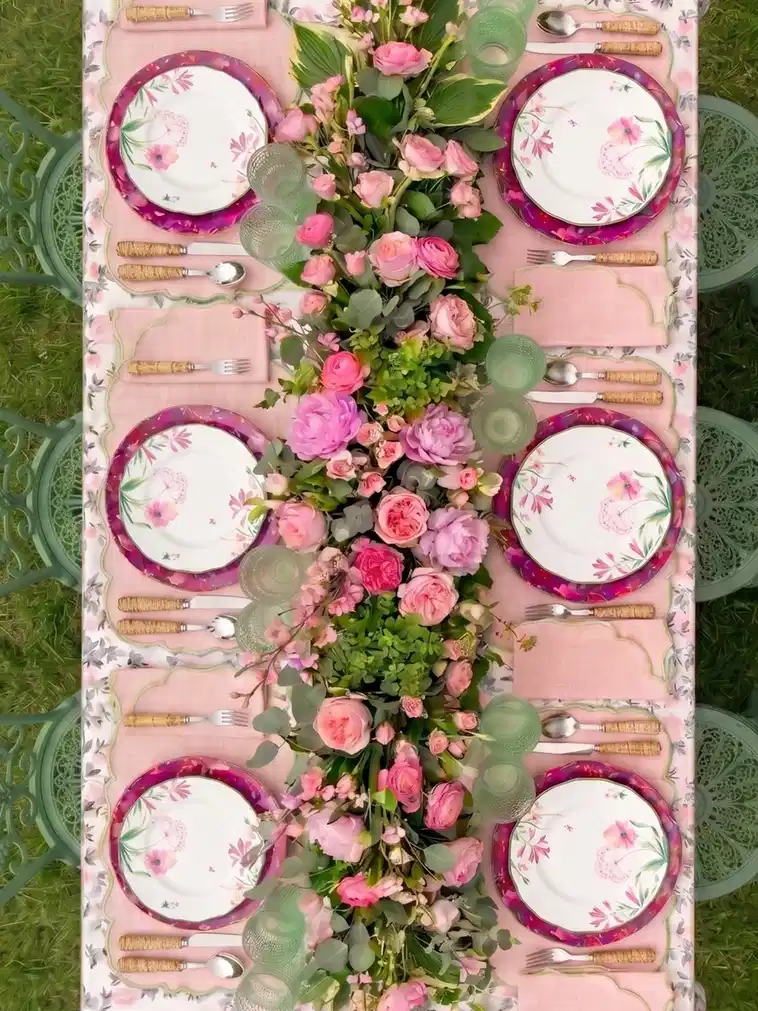 Overhead view of outdoor table settings with pink flowers and green foliage arranged between plates