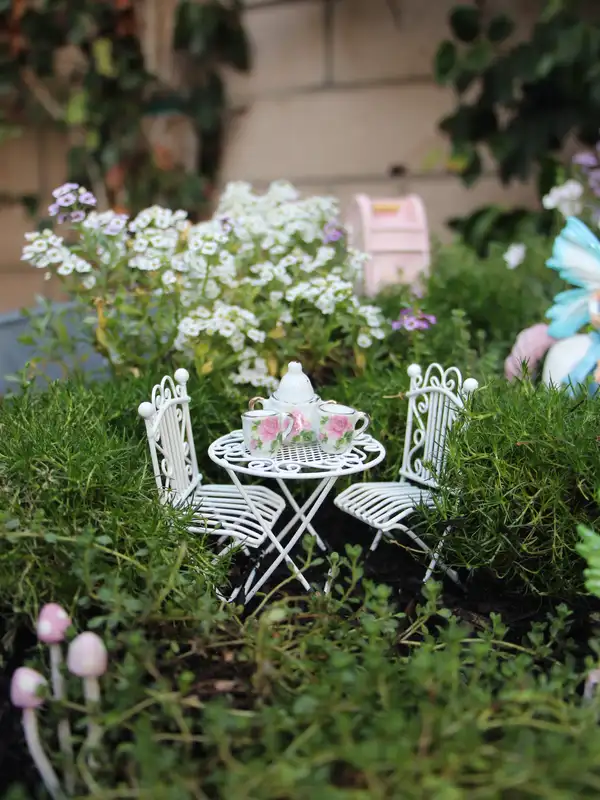 Small fairy garden with white miniature table and chairs surrounded by pale flowers and greenery