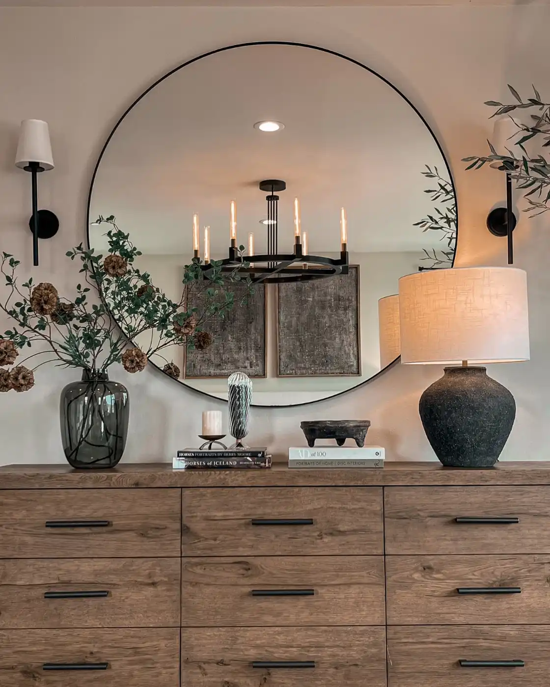 Long dresser beneath large round mirror with leafy branches, table lamp, books, bowl, and wall sconces