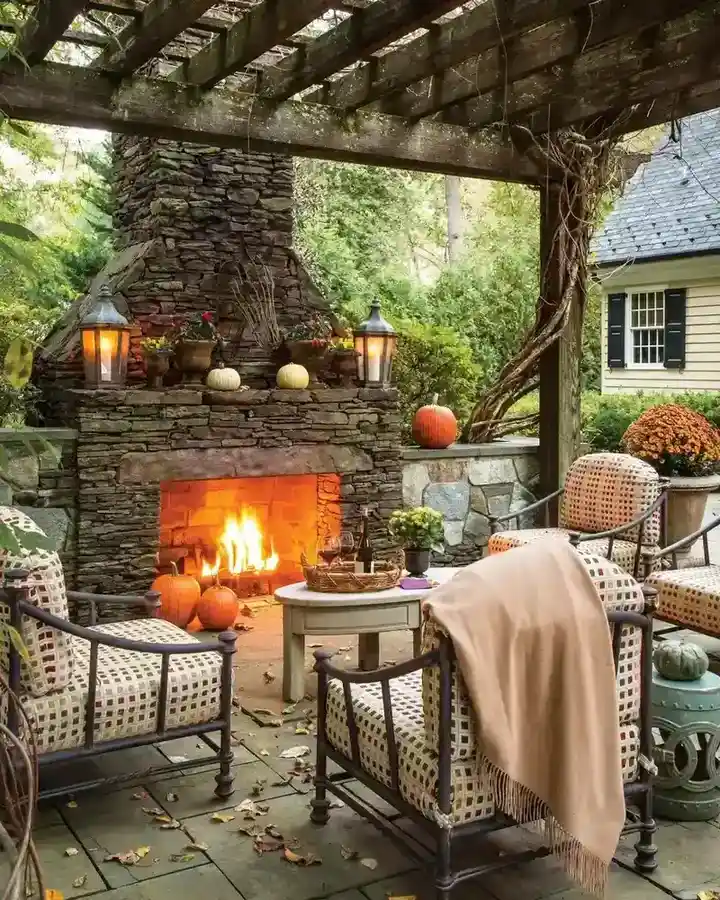 Rustic covered porch with a stone fireplace, fire, chairs, wood pergola ceiling, blankets, and pumpkins.