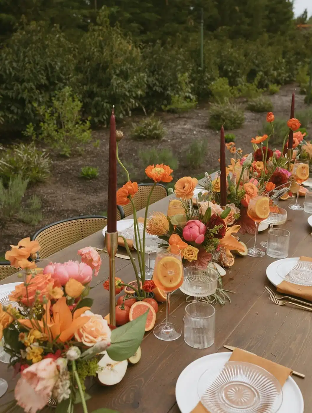 Outdoor dinner table with peach and coral flower arrangements, slim candle holders, and pale tableware