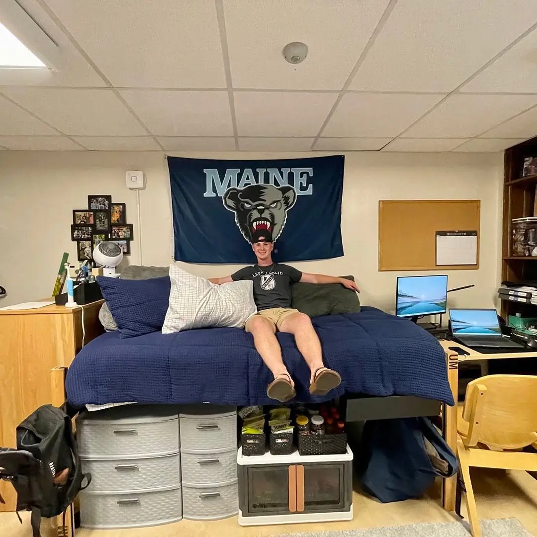 Raised dorm bed with navy bedding, a large Maine flag, drawer units, bins, and storage underneath