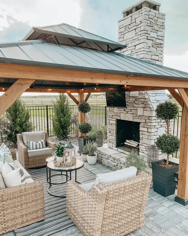 Covered porch with stone fireplace, wicker chairs, pale cushions, wood ceiling, and greenery