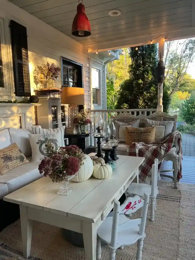 Covered dining porch with a long light wood table, chairs, hanging lights, decorative details, and plants.