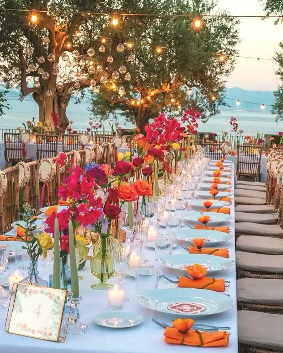 Festive outdoor dining table decorated with colorful flowers, warm string lights, and coordinated place settings