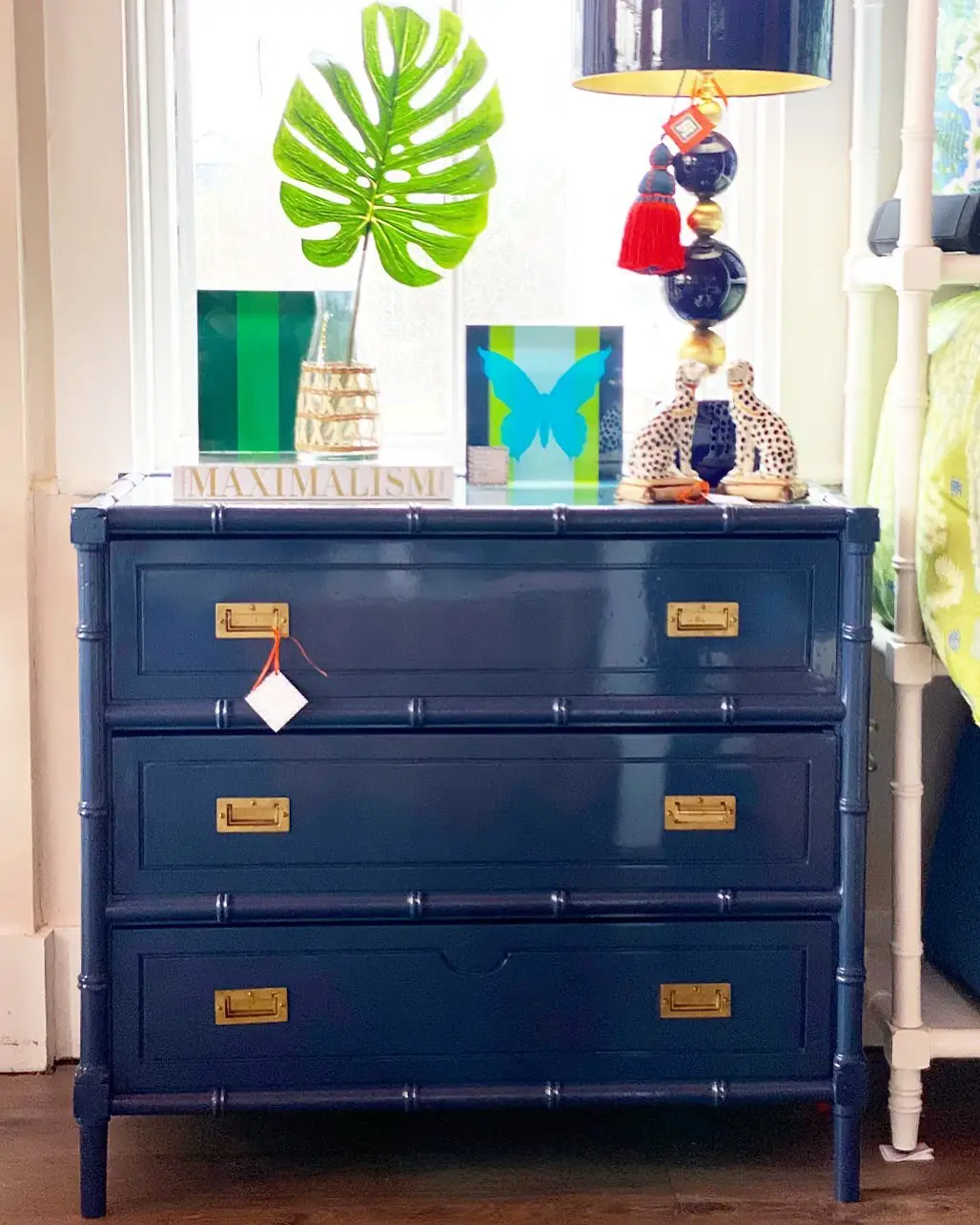 Navy dresser with colorful framed art, large green leaf, stacked books, and playful table lamp