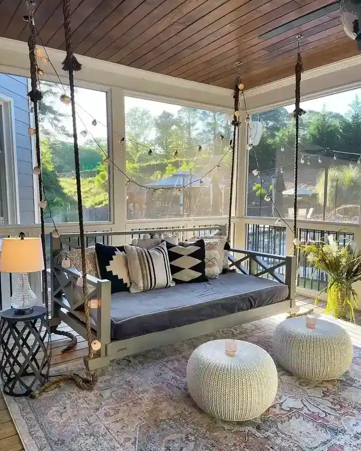 Screened porch with wood ceiling, sofa, dark patterned rug, two round ottomans, and open windows.