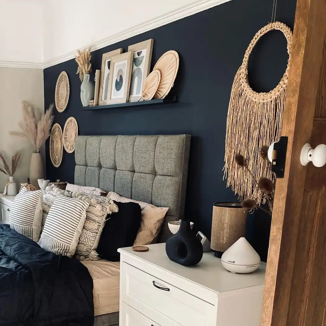 Black accent wall bedroom with beige woven wall decor, gray upholstered headboard, navy bedding, striped pillows, and natural wood accents