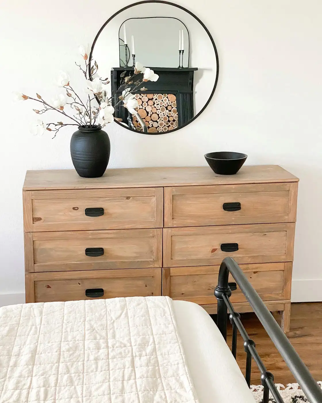Natural wood dresser beneath round mirror with tall dark vase of branches and low black bowl