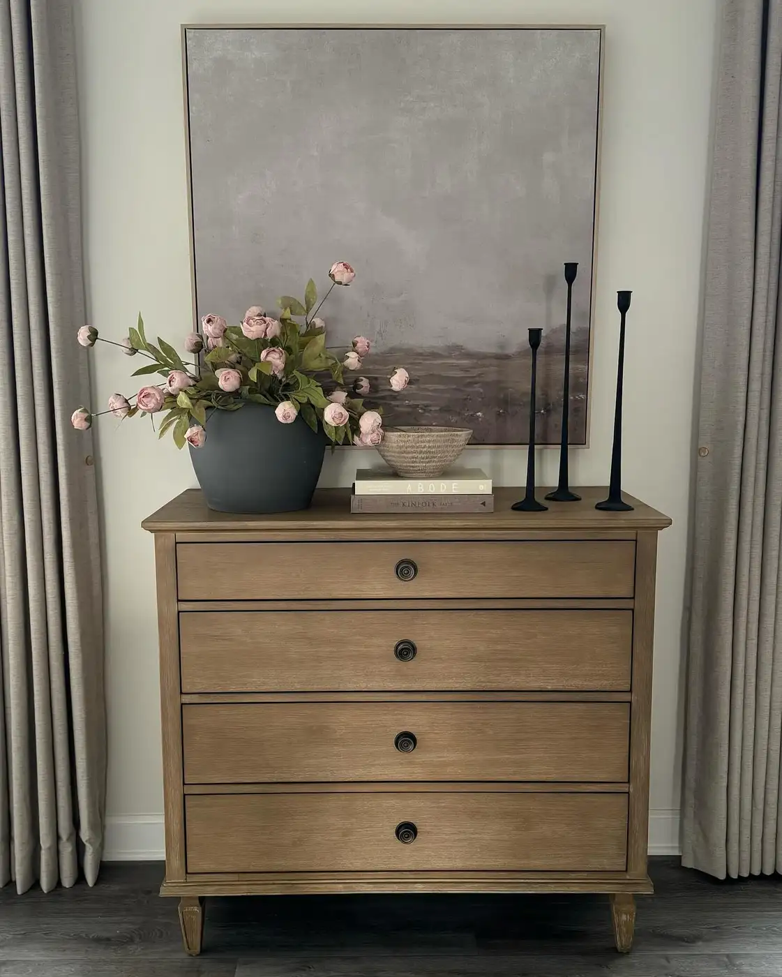 Wood dresser beneath oversized artwork with pink flowers, black candlesticks, stacked books, and shallow bowl